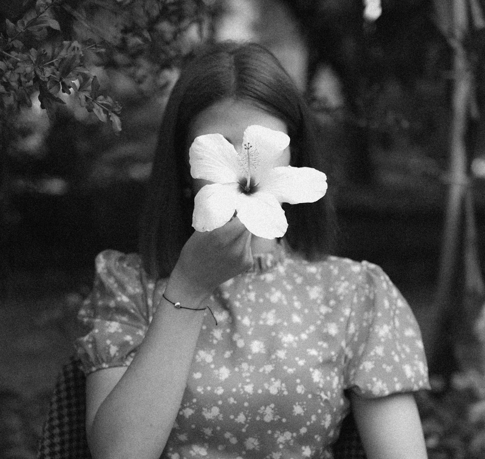 A woman wearing a floral dress and holding a lily in front of her head.