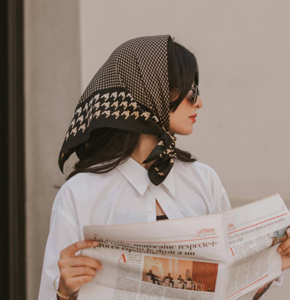 A woman with a black and white bandana wearing sunglasses and holding a newspaper.