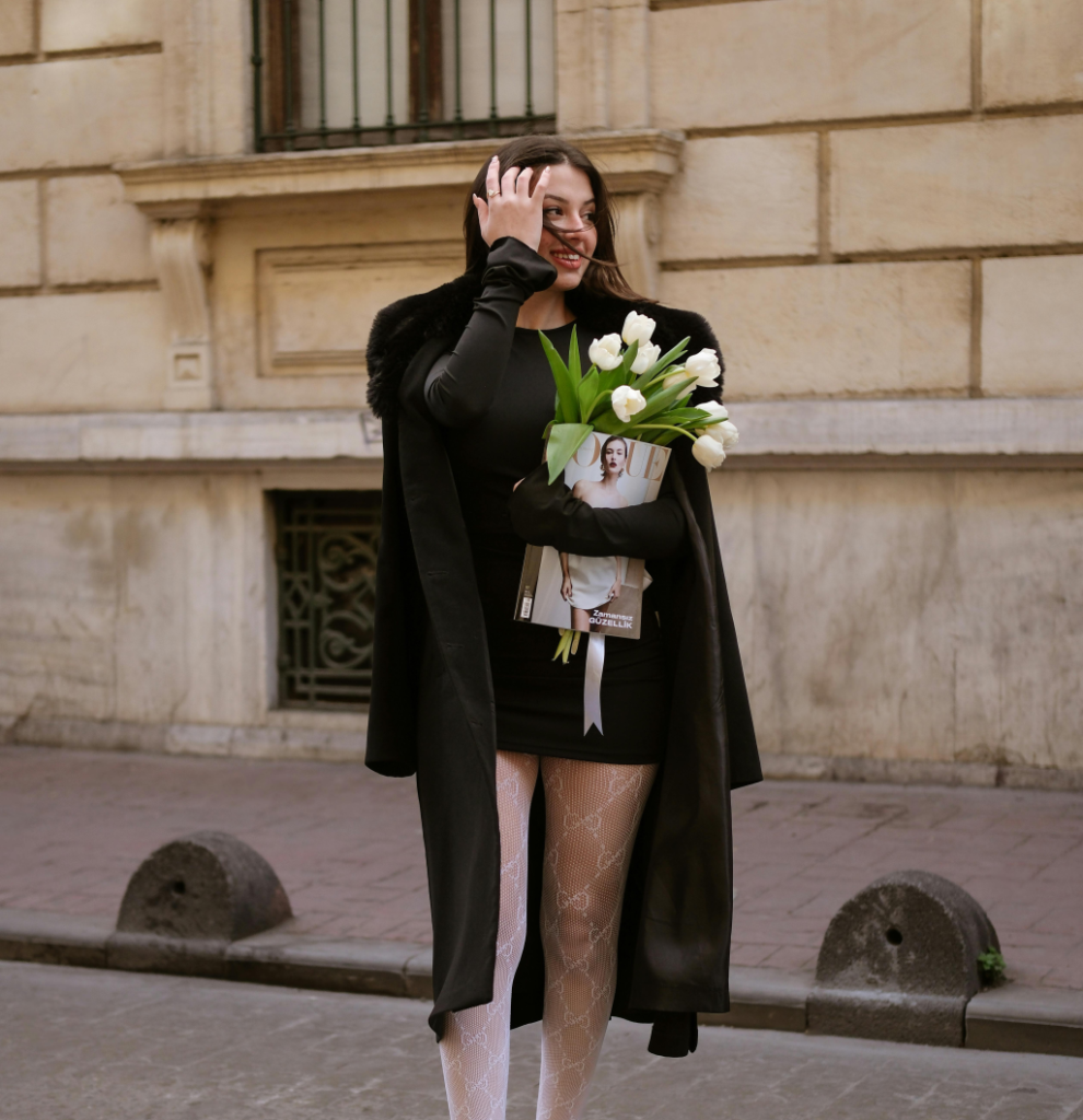 A woman with a black dress and a black coat holding white flowers.