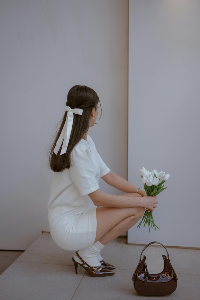 A woman in white with brown heels and a brown handbag is holding white flowers.