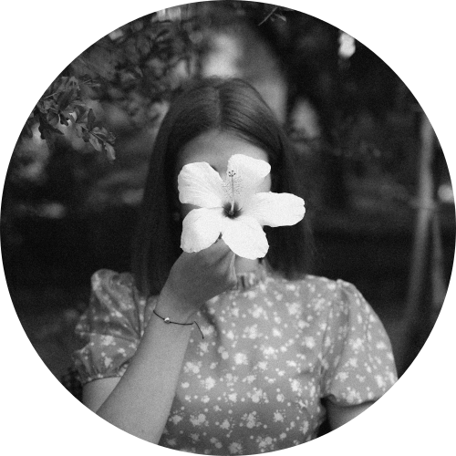 A woman wearing a floral dress and holding a lily in front of her head.